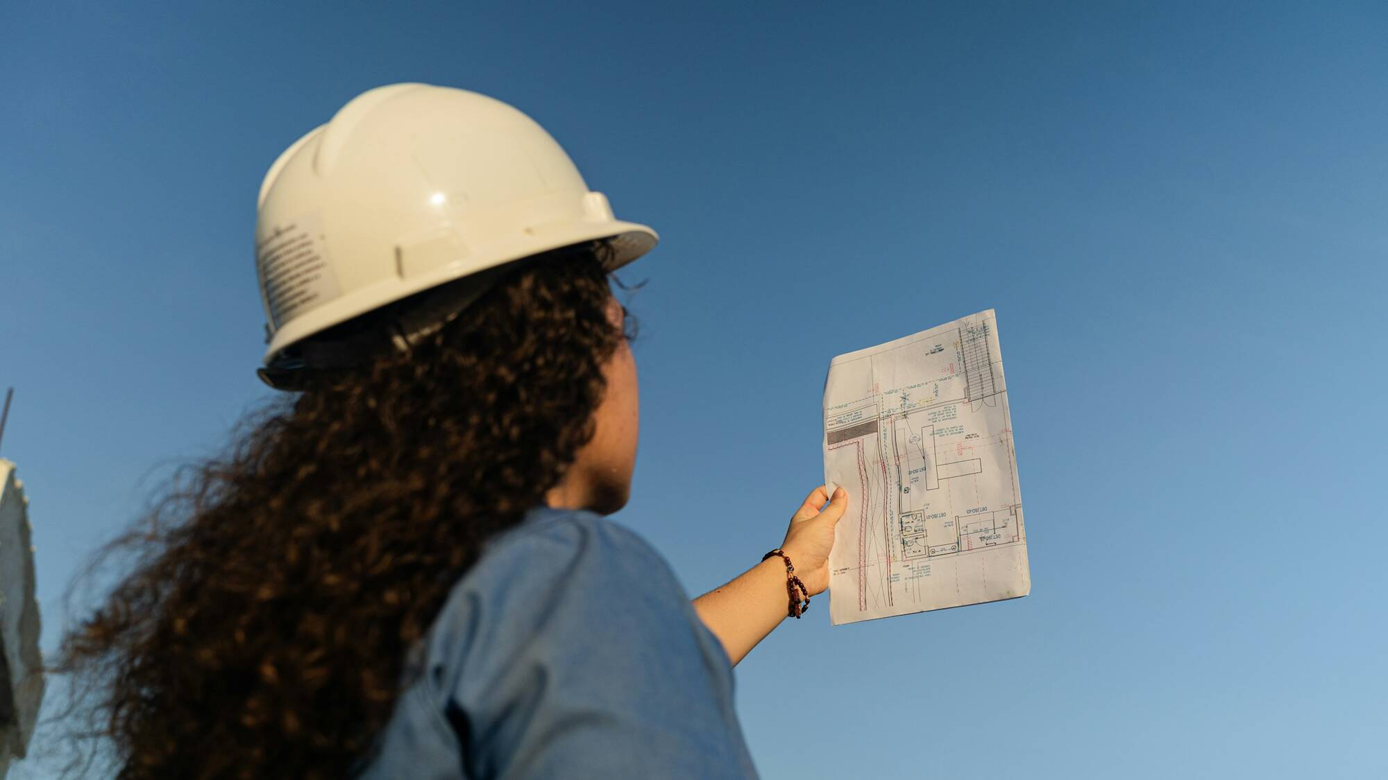 Persoană cu cască albă de protecție care ridică un plan împotriva unui cer albastru